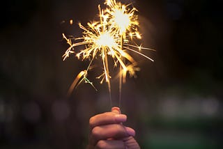 A hand holds two lit sparklers on a dark, blurry background