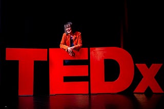 The author Trisha Lewis on the TEDx stage. Leaning over the ‘E’ of the giant red TEDx blocks.
