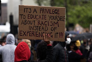 Protestor holding a cardboard sign that reads “It’s a privilege to educate yourself about racism instead of experiencing it!”