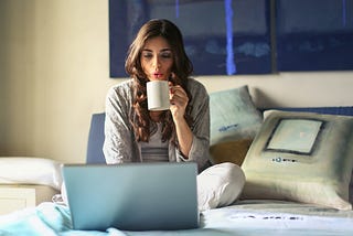 woman sitting on bed on laptop blowing on a cup of coffee