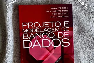 Imagem da capa do livro ‘Projeto e Modelagem de Banco de Dados’, segunda edição, publicado pela Elsevier. A capa tem um fundo vermelho escuro com elementos geométricos abstratos em tons de vermelho mais claro. Os nomes dos autores aparecem no topo da capa em letras brancas: Toby Teorey, Sam Lightstone, Tom Nadeau e H.V. Jagadish. O título do livro está em letras grandes e brancas no centro da capa. No canto inferior esquerdo está o logotipo da editora Elsevier.