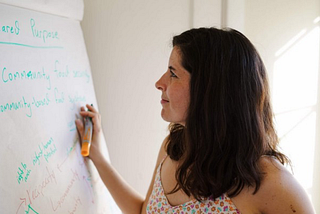 A white woman with shoulder-length brown hair wearing a flower-print camisole holds an orange marker and gazes at a white flip chart with indeterminate green writing.