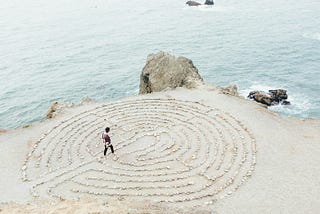 An image of a woman walking on the beach into a maze made of stones illustrates the uncertainty we often face about whether to act immediately when confronted with a challenge or barrier. By taking time to pause and avoid reacting impulsively, we practice presence and approach these situations with greater clarity and intention.