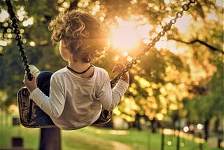 rear view of young boy with curly brown hair wearing long sleeve white top mid-air on a swing over grassy lawn