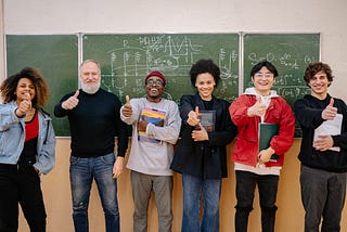 Students and a teacher standing in front of a blackboard with their thumbs up.