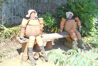 Photograph of two characters made out of flower pots, seated on a bench in an English country garden and made to resemble Bill and Ben, The Flowerpotmen who featured on a 1950s BBC TV programme called ‘Watch With Mother’.