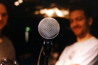A close-up of a microphone, with two blurry listeners in the background.