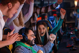 Three adults cheering on a fourth adult in front of a computer with headphones on, presumably cheering him on winning a video game tournament from the article Why Fun is Essential for Survival and Liberation by Gem Blackthorn