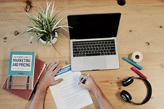 An overhead shot of a laptop, a pair of hands writing on a piece of paper, a book, a plant, headphones pens and tape.