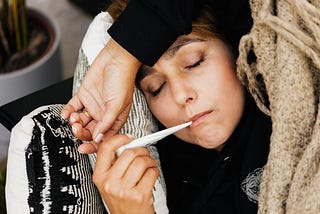 A woman holds a thermometer in her mouth as she closes her eyes in bed.