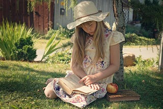 A young girl reading a book by a tree.