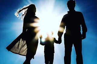 A mother and father holding the hands of a small child, the sun is in front of them creating a silhouette of their figures.