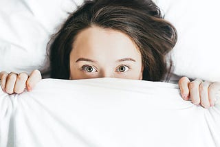 A scared woman lying on bed covered with a white blanket with only her eyes and forehead exposed