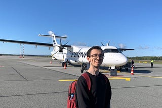 A young man stands in front of a Finnair propeller plane on an empty runway. He wears a red backpack and smiles slightly, framed by clear skies and open tarmac.