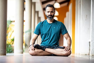 A man sits cross-legged in a meditative posture on the floor of a bright corridor, wearing a teal athletic shirt and black socks. He appears calm and focused, with natural light highlighting the peaceful setting. Reflecting the perspective of Ramneek Bhogal on integrating wellness into busy professional lives.