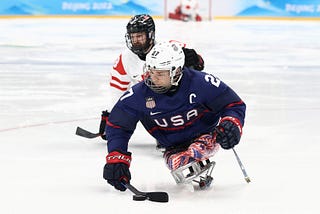 Josh Pauls is a white man wearing a white helmet with a metal face cage and a navy blue hockey jersey with USA and a letter C indicating that he is the team captain on the wrong. The number 27 is written on the shoulder. He is sitting in a sled hockey sled with a carbon fiber casing cpvering his lower body decorated to look like an American flag. He is a double above the knee amputee. He is holding two sticks, and using one to control the puck. A player from Canada is in the background.