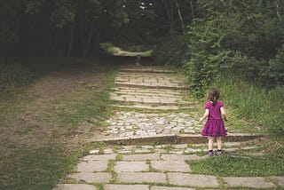 Little girl on a stone path, looking after someone she cares for — far off in the shadows.