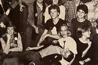 According to ChatGPT, this is a black-and-white photo of a group of high school students posing together, likely from the 1980s. They are arranged in several casual rows, smiling and leaning against each other, with one student lying playfully across the front. The scene conveys youthful camaraderie and nostalgia.