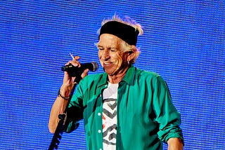 Keith Richards in a green shirt and black headband holding a microphone (and a cigarette) in front of a blue lightboard