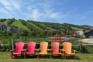 Five Muskoka chairs (2 red, 3 orange) lined up against the backdrop of a ski hill and blue skies in the summer.