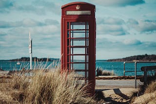 A red telephone booth standing empty in front of the shore.