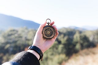 A person’s arm is outstretched and is holding a compass in front of a scenic view of sand, mountains, and shrubbery in the backgound.