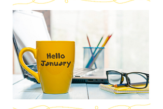 A yellow “Hello January” mug, computer, notebook, pencils, and a pair of glasses sit on a table in a bright and sunny room.