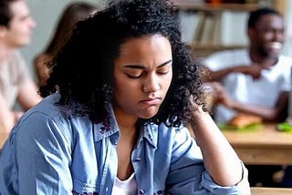 In focus is a person sitting alone with a sad expression with a background of a group of people talking and laughing.