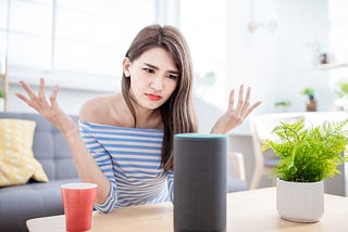 A woman sitting in a living room looks at her smart speaker with disdain and raises her hands in frustration.
