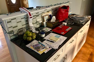 Author’s photo of a kitchen counter with piles of papers, a laptop, red purse, basket of apples