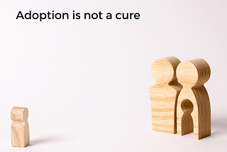 Text reads, “adoption is not a cure” and two wooden figurines are standing side to side. In one, there’s a hole in the shape of a small child. The child figurine is standing apart from the two ‘adults’.