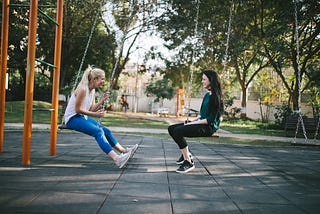 Two women sitting on swings, chatting and laughing.