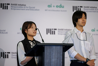 Two people stand on stage presenting their work. In the background is a white board featuring the MIT RAISE and ai + edu logos.