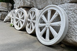 Wheel of Buddhist Dharma, a symbol of teaching in Buddhism. White marble wheels are set in a row, leaning against large boulders, with a statue of a white stork in the background. Installation at a Buddhist temple. Image credit: Oxana via Adobe Stock.