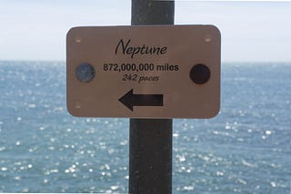 A small signpost with the sea in the background, reading ‘Neptune: 872,000,000 miles, 242 paces’.