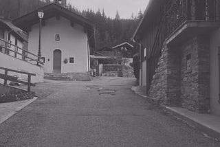 Looking uphill on a narrow paved road. Lampost and small church door on left, stonework on right. Black and white image. Trees in background