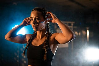 A muscular girl with long braids is shown from the chest up, adjusting her headphones and looking determined before starting her set