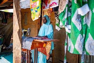 A woman works outside her home at a sewing machine table surrounded by a variety of colorful material hanging overhead and behind her.