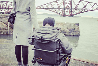 Anna and her brother pictured overlooking a bridge in South Queensferry. They are both wearing grey coats, and her brother sits in an electric wheelchair