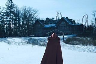 Person in cape with back to viewer, facing snowy yard and wooden house with windmills behind it.