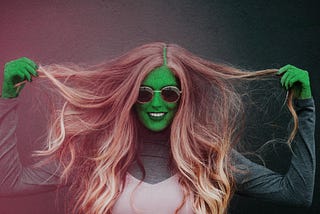 Green-skinned woman with cracked skin effect wearing a long-sleeved sweater and pink tank top, staring intensely at the camera — Halloween makeup portrait.