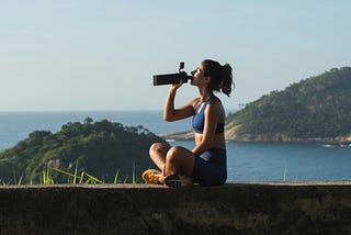 A person with long hair tied back sits cross-legged on a stone ledge outdoors, wearing a blue athletic top, matching shorts, and yellow shoes. They are holding a large water bottle up to their mouth as if drinking. In the background, there are green hills and blue water under a clear sky.