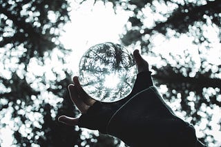 A hand holds a glass ball in a forest underneath some trees