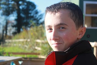 The author as a young child. Approximately 10 years old. He has short, brown hair and the right half of his face is in shadow. He has a smile with pursed lips. He’s wearing a red and brown jacket. In the background is a small hill of grass, tall trees, and a blue sky.