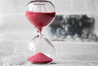 Clear glass hourglass with red sand running through it against a greyed out backdrop of printed paper.