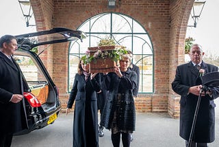 A wicker coffin garlanded with flowers being carried into a funeral service by four pallbearers. Only the two at the front are visible: a middle-aged woman and a younger women who might be her daughter.