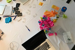 Picture of a table with crumpled notes, a computer, pens on top a white tablecloth.