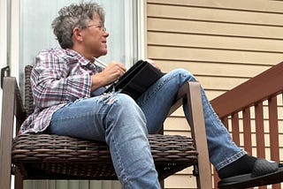 Photo of an older woman with short gray hair before she had it colored sitting in a deck chair with an iPad and Apple pencil watching the neighbor’s dog which you can’t see but you can see she’s wearing a Nike slide and dangling her foot because she still likes to think of herself as casually childlike