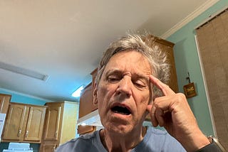 An older man with gray hair is indoors, He appears to be yawning and is sitting in a kitchen with wooden cabinets and a light blue wall.
