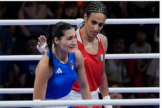 Image shows two women. In blue, looking tearful, Italian boxer, Angela Carini; in red, Algerian boxer Imane Khelif. Imane is reaching out to console Angela.
